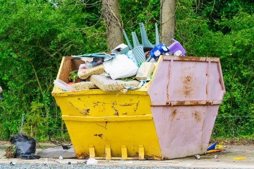 A large yellow and pink industrial skip bin filled with assorted garden and household rubbish, including plastic containers, foam padding, and fabric materials, situated outdoors on a gravel surface near dense green foliage and trees, with some smaller waste items and black plastic bags on the ground nearby, representing a typical scene for rubbish collection or clearance services in Upminster. The skip shows signs of rust and wear, indicating it has been used for collecting waste from garden clearance or domestic decluttering projects, with a mix of textures and materials visible, such as the smooth plastic and rough fabric. The setting suggests a residential or semi-rural environment, relevant to Garden Clearance Upminster’s services for removing rubbish from local gardens and properties.