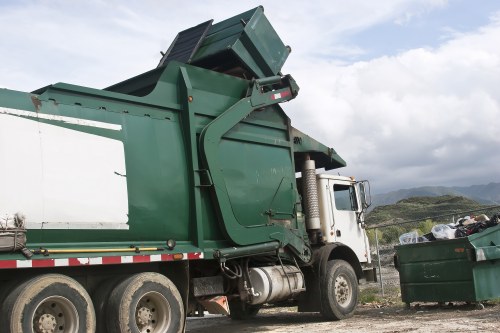 A large green waste collection truck with a white cab is parked outdoors on a gravel surface, with its rear compartment raised slightly for loading. The truck's metallic green body shows signs of use with some dirt and scratches, and the hydraulic arm and lifting mechanism are visible at the back. To the right of the truck, there is a green wheelie bin filled with mixed rubbish, including plastic bags, paper, and miscellaneous debris. The background features a partly cloudy sky with patches of blue and distant rolling hills, suggesting a rural or suburban environment. The scene relates to rubbish removal services in the Upminster area, and the presence of the waste truck and bin reflects the typical equipment used for garden and general waste clearance, consistent with services provided by Garden Clearance Upminster.