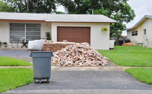 A residential driveway in front of a single-story house with a white exterior and brown garage door. In the center of the driveway, there is a large pile of light-colored bricks and rubble, likely from a recent garden or property clearance. To the left of the pile, a dark grey wheelie bin is positioned on the black asphalt surface. The house has a large front window partially covered by blinds, and a small potted plant is visible near the window. The surrounding area includes neatly maintained green lawns on both sides of the driveway, with a concrete path leading to the house entrance. Mature trees are visible in the background, along with a neighboring property to the right. The scene suggests a professional rubbish clearance service undertaken by Garden Clearance Upminster, located in the Upminster area, providing efficient removal of construction debris associated with garden or property renovation projects.