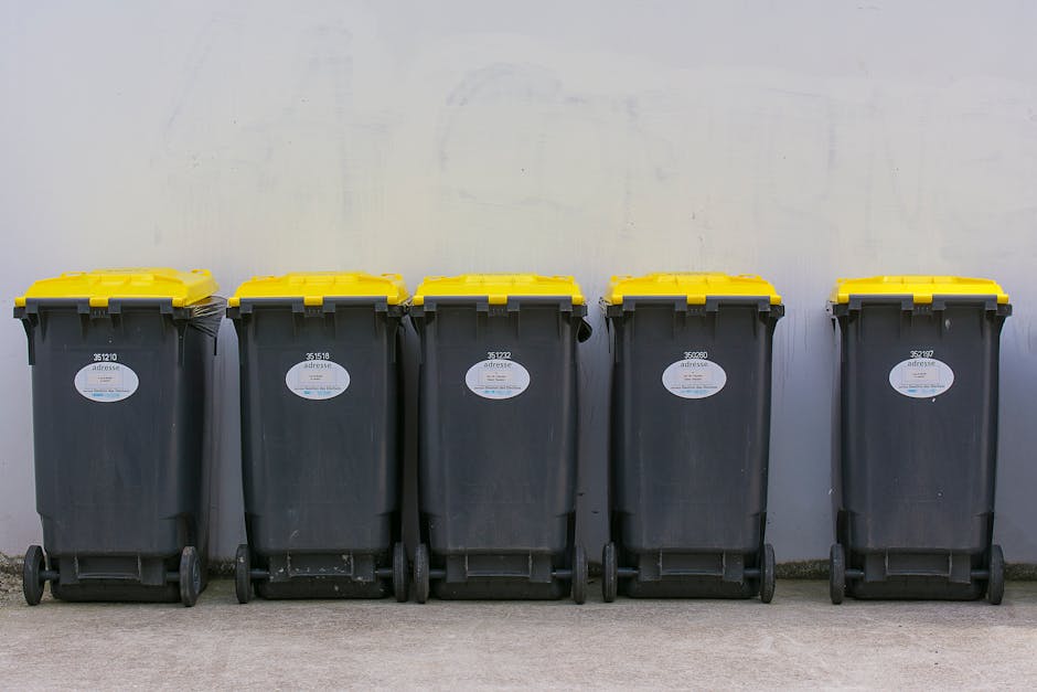 A white rubbish collection truck is parked on a narrow cobblestone street beside a row of older, multi-story residential buildings with worn facades and small windows. The truck's rear compartment is 