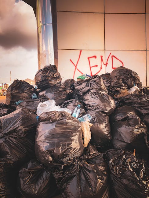 A close-up view of a pile of mixed rubbish and waste materials situated on a gravel surface beside a weathered concrete curb. The waste includes black and transparent plastic garbage bags, some of whi