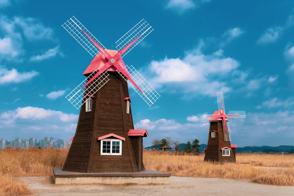 A traditional black windmill with four large lattice-style sails stands in an open grassy landscape under a clear blue sky. The windmill's cylindrical base is made of dark painted wood, with small win